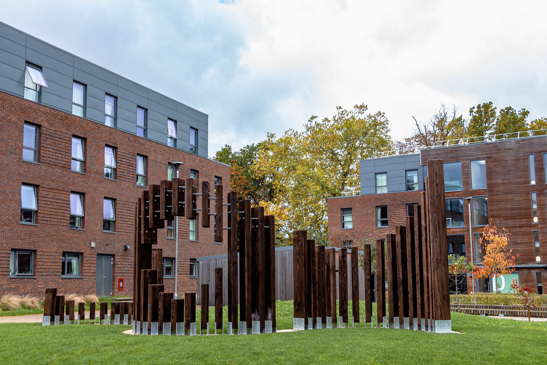 A wooden sculpture consisting of vertical beams in a winding shape, on top of a small grass mound with halls of residence in the background